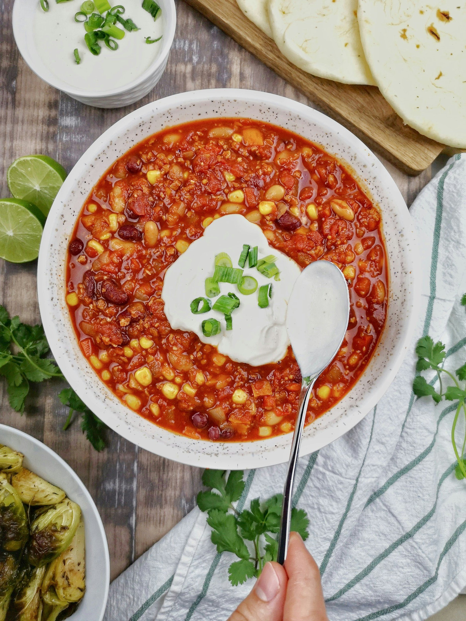 Bowl of hearty chili topped with sour cream and green onions, served with lime wedges and a side of roasted Brussels sprouts on a wooden table.