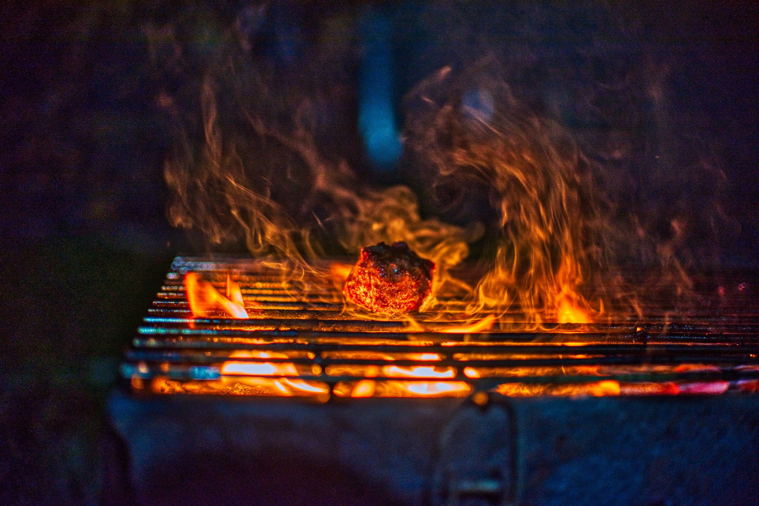 Grilled meat sizzling over an open flame, with smoke rising in a dark outdoor setting. The image captures the vibrant colors of the fire and the texture of the meat on the grill, evoking a sense of outdoor cooking and barbecue.
