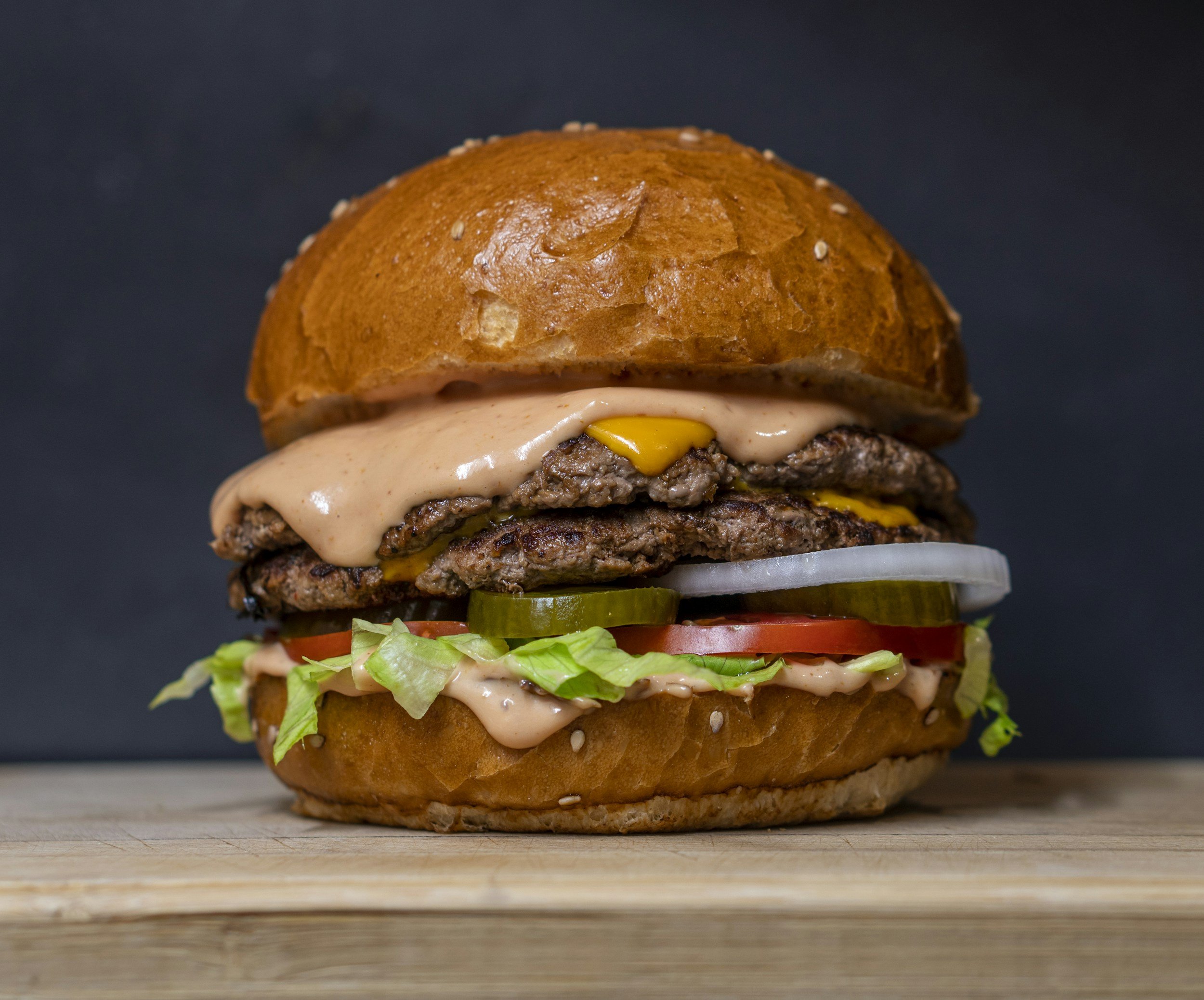 Juicy double cheeseburger with melted cheddar cheese, special sauce, lettuce, tomato, pickles, and onions on a sesame seed bun, set against a dark background.